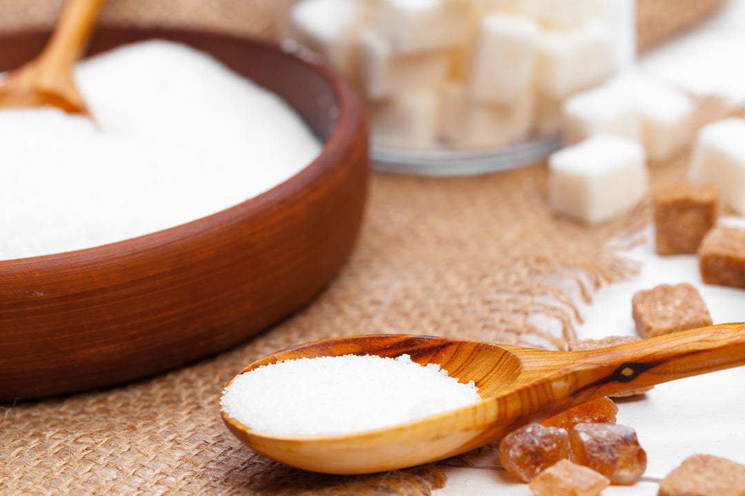 A close-up of a wooden spoon filled with fine white granulated sugar, with a rustic wooden bowl and raw sugar cubes out of focus in the background on a burlap surface.